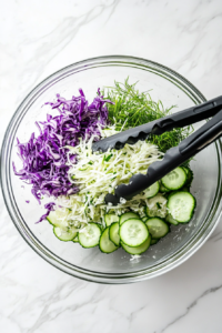 The cabbage, cucumber, and green onion salad is being tossed with the sunflower oil, vinegar, and salt dressing inside a large clear glass mixing bowl. A pair of black salad tongs is being used for even mixing. The scene sits on a white marble countertop, and the vibrant colors blend together, making the salad look fresh and appetizing.