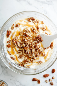 Top-down view of caramel dip singles being gently folded into the cheese and pecan mixture using a white spatula in a clear glass bowl. The creamy beige mixture is marbled with golden caramel swirls, enhancing its sweetness. The ingredients are being incorporated evenly to ensure every bite has the perfect blend of creamy and caramel flavors.