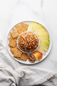 Top-down view of the fully assembled caramel pecan cheeseball served with sliced apples, graham crackers, and pretzel chips on a white ceramic plate. The cheeseball is coated in pecans and drizzled with caramel, making it an ideal sweet and salty appetizer or dessert dip. The clean white marble background enhances the simple yet elegant presentation.