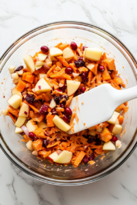 Top-down view of the complete salad being gently tossed in a clear glass bowl using a white silicone spatula. All ingredients—grated carrots, diced apple, dried cranberries, chopped onion, and creamy dressing—are evenly mixed. The bowl rests on a clean white marble counter, presenting a fresh, colorful, and well-blended salad.
