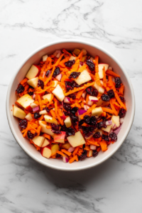 Top-down view of a fresh carrot apple salad served in a white bowl on a white marble countertop. The salad includes coarsely grated carrots, diced red apples, dried cranberries, and finely chopped purple onion all tossed in a creamy mayo dressing. This colorful, crunchy salad is simple, vibrant, and full of texture and flavor.