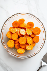 Freshly peeled and cut carrots are tossed with olive oil kosher salt and black pepper in a large clear glass bowl. The seasoning evenly coats the carrots, preparing them for roasting. The simple white marble countertop and consistent use of kitchen tools maintain a clean and focused cooking environment that’s easy to follow.