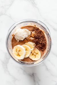 This top-down image shows the blended smoothie being checked for consistency. A small measuring cup of almond milk is placed nearby, ready to be added if needed. The smoothie inside the blender appears thick and creamy, and everything is set on a white marble countertop, keeping the kitchen scene realistic and clean.