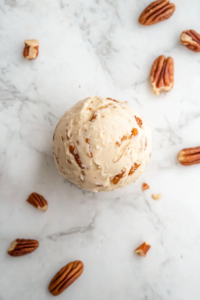 Top-down view of hands shaping the creamy caramel and pecan mixture into a ball over a white marble countertop. The soft mixture is formed into a round shape and placed on a sheet of clear plastic wrap. The rich texture of the mixture is visible, and it’s ready to be tightly wrapped and chilled for setting and flavor development.