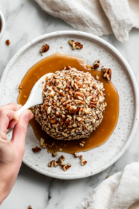 Top-down view of the chilled caramel pecan cheeseball being rolled in a generous layer of finely chopped toasted pecans on a white ceramic plate. A spoon drizzles extra caramel sauce over the top, adding a glossy, sweet finish. The setup on the white marble countertop highlights the contrast between the nutty coating and creamy interior.