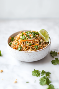 A top-down shot of a chilled soba noodle salad beautifully arranged in a white ceramic bowl, sitting on a clean white marble countertop. The noodles are mixed with finely grated carrots, fresh chopped cilantro, and sprinkled with coarsely chopped salted peanuts. A lime wedge is placed at the side, enhancing the fresh, zesty appearance of this vibrant and healthy salad.