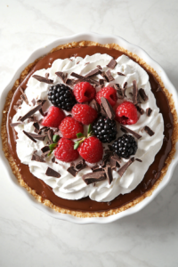 Top-down view of a rich chocolate no-bake cheesecake beautifully set in a white 9-inch ceramic pie dish. The cheesecake is garnished with fluffy whipped cream, chocolate shavings, and fresh berries. It rests on a clean white marble countertop, with a smooth chocolate surface and golden graham cracker crust, creating an elegant yet indulgent dessert presentation.