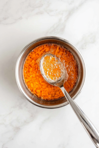Coarsely chopped carrots are boiling in a medium stainless steel saucepan on a white marble countertop. The carrots are softened to create the base of the soufflé. Once tender, they will be drained and slightly cooled before blending. This step brings out the natural sweetness and vibrant color of the carrots.