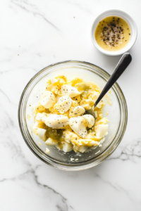 Chopped boiled eggs are added to the prepared creamy dressing in a clear glass bowl and are being gently stirred with a silver spoon. This step ensures each chunk of egg is coated with rich, tangy flavor. The mix reveals a well-blended texture that’s ideal for sandwiches, crackers, or a lettuce wrap.