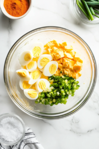 A clear glass bowl sits on a white marble surface, filled with chopped boiled eggs, mayonnaise, yellow mustard, green onions, paprika, salt, and pepper. This image shows all the ingredients before mixing, capturing the vibrant colors and textures that will come together to make a flavorful and creamy egg salad.