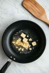 Finely chopped garlic is sizzling in fully melted butter inside a black skillet on a white marble surface. The garlic is turning golden and releasing its fragrance, creating a delicious aroma. This step lays the essential flavor foundation for the mushroom dish. A wooden spatula rests beside the skillet for stirring.
