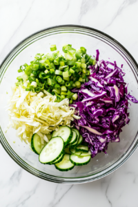 Freshly chopped green onions are being added into a clear glass bowl that already contains shredded cabbage and cucumber slices. The green onion adds a sharp flavor and a vibrant green color to the salad. The bowl sits neatly on a white marble kitchen countertop, with a cutting board and a sharp knife nearby for prep work.