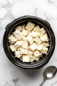 A top-down view shows coarsely chopped white chocolate chunks placed at the bottom of a black crock pot sitting on a clean white marble countertop. The white chocolate is the base layer for this dessert recipe and is ready to melt slowly, setting the foundation for a rich and creamy chocolate mixture.