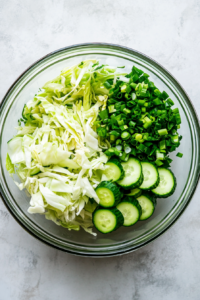 A top-down view of green onions being chopped and added to a clear glass bowl that already contains finely sliced cabbage and cucumber. The clean white marble background enhances the fresh green color of the onions, which contribute a mild, oniony flavor and vibrant color contrast to the salad mix.