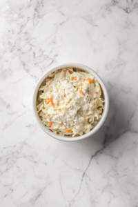 Top-down view of a bowl filled with creamy coleslaw made from chopped green cabbage, shredded carrots, and minced onion. The vegetables are coated in a rich dressing made from mayonnaise, vinegar, mustard, sugar, and pickles. The dish is served chilled in a white bowl over a clean marble background, capturing freshness and texture.