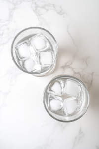 Several clear glasses are neatly filled with fresh ice cubes, sitting atop a white marble countertop. The glasses are ready to receive the chilled Brazilian lemonade. This step is essential for serving the drink refreshingly cold and enhancing the overall experience of the creamy, citrusy beverage during hot or sunny weather.