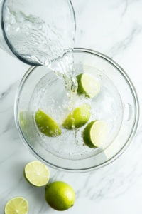 Cool, clear water is being poured from a glass measuring cup into a blender already filled with lime wedges. The water begins to surround the citrus pieces as preparation for blending begins. This step ensures the lime flavor infuses evenly into the liquid base for a well-balanced and fresh Brazilian lemonade.