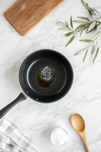 A clean top-down shot of a black saucepan placed on a white marble countertop with coconut oil melting inside. The oil is clear and glossy, signaling it’s heating properly over medium heat. This is the starting step to prepare a vibrant cranberry jalapeño dip, where each layer of flavor begins in this simple preparation.