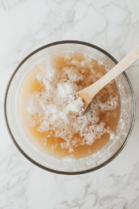 A transparent glass mixing bowl sits on a white marble countertop, containing a combination of warm water, dissolved yeast, white sugar, soft lard, fine salt, and gradually added all-purpose flour. A wooden spoon is being used to mix the ingredients, forming a shaggy dough. This step ensures the dough is properly combined and develops gluten for a smooth and elastic texture.