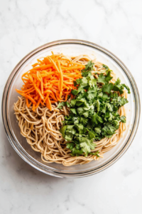 This top-down image shows a large clear glass bowl placed on a white marble countertop filled with cooked soba noodles, finely grated carrots, and freshly chopped cilantro. The ingredients are layered together before tossing. The setup is bright and clean, capturing the step where the vibrant vegetables are mixed with the earthy noodles for a colorful and healthy dish.