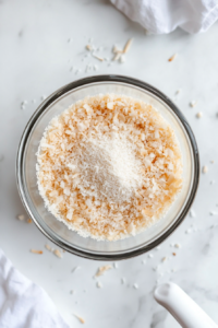 A top-down shot shows a glass mixing bowl on a white marble surface containing spelt flour, unsweetened desiccated coconut, baking powder, and salt. A white spatula or whisk is used to blend the dry ingredients. The scene is kept minimal and clean to focus on preparing the dry base mixture for the cake batter.