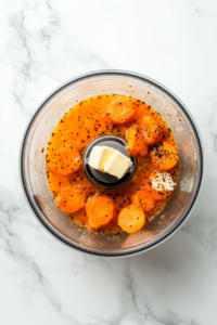 Cooked carrots are being blended with melted butter and vanilla extract in a clear glass food processor. The smooth mixture is bright orange in color and forms the creamy base of the soufflé. This step creates a rich, aromatic carrot purée that will give the dish both texture and flavor.