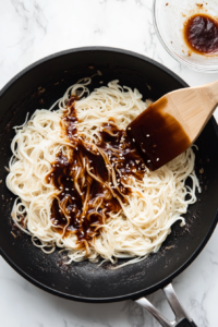 Cooked noodles are added to the skillet over sautéed aromatics while the soy-teriyaki sauce mixture is being poured in. The ingredients blend together in a black skillet on a white marble cooktop. The noodles are starting to absorb the savory, slightly sweet sauce, preparing them to be fully stir-fried in the next step.