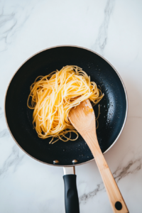 A top-down image of cooked spaghetti noodles being added to a black nonstick pan with hot olive oil. The noodles are soft and lightly glossy, being gently tossed with a wooden spoon. This step starts the stir-fry process that builds the base for the flavorful teriyaki noodle dish.
