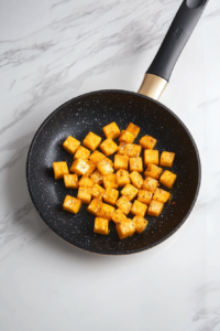 Diced pumpkin is added to the sautéed onions and garlic in the black skillet. The golden cubes are beginning to soften and release their natural sweetness. This step allows the pumpkin to absorb the savory base flavors. The white marble countertop keeps the scene clean, with visible ingredients ready for the next stage of cooking.