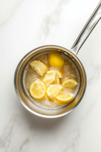 The stainless steel saucepan now contains a lemon-infused syrup that has been removed from heat and allowed to cool for 15–20 minutes. The diced lemon peel floats gently in the pale yellow liquid, and the condensation has faded, indicating the syrup is ready for the next step in the lemon sorbet preparation.