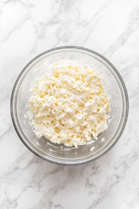 The mixed coleslaw is now resting in a large glass bowl covered with a clear airtight lid, placed on a clean white marble countertop. This step is crucial for allowing the flavors of the creamy dressing to blend with the fresh vegetables, enhancing the overall taste and texture of the coleslaw as it chills.