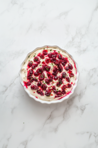 Top-down view of the finished cranberry jalapeño cream cheese dip presented on a white ceramic dish over a white marble countertop. The deep red cranberry topping with visible jalapeño specks is layered over creamy white cream cheese. It’s beautifully chilled and served with crispy crackers, making it a perfect party appetizer with bold flavors.