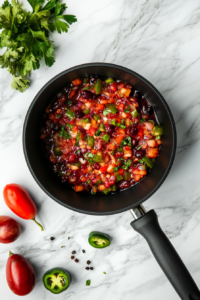 A top-down view of the just-cooked cranberry jalapeño mixture resting in a black saucepan atop a white marble countertop. The vibrant red mixture is cooling, steam gently rising, allowing the flavors to settle. This important step prepares the topping for blending or serving, ensuring it won’t melt the cream cheese layer beneath.