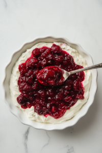 A top-down image showing the vibrant red cranberry jalapeño mixture being carefully spooned over a smooth cream cheese layer on a white ceramic plate. The layers contrast beautifully. This final assembly step is key before chilling, ensuring the spicy-sweet mixture fully infuses into the creamy base for optimal flavor.
