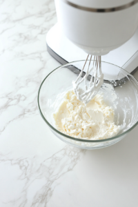 A mixing bowl shows butter and erythritol being creamed with a white electric mixer until light and fluffy. Next to it, cracked eggs, plain yogurt, and vanilla extract in small glass bowls are ready to be added. The countertop is white marble, keeping the scene consistent and emphasizing the wet ingredient preparation stage.