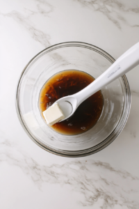A clear glass mixing bowl contains softened unsalted butter, monk fruit sweetener, and a spoonful of blackstrap molasses being creamed together using a white electric hand mixer. The bowl is placed over a white marble countertop. This step combines fats and sweeteners to create a light and fluffy base for the carrot cake batter.