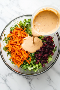 Top-down view showing the creamy dressing made from mayo, lemon juice, sugar, salt, and pepper being poured into the salad mixture containing grated carrots, apples, cranberries, and onions. The glass mixing bowl is placed on a white marble counter. This step adds creaminess and flavor, coating the vibrant salad ingredients.
