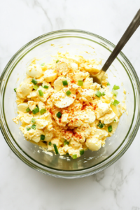 This image shows the completed mixture of creamy egg salad in a clear glass bowl, with the ingredients stirred until fully combined. You can see the chopped green onions, specks of paprika, and creamy texture. Placed on a white marble countertop, this step confirms the salad is ready to serve or chill.