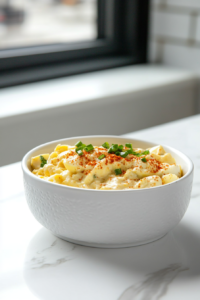 A top-down image showing a creamy homemade egg salad served in a white ceramic bowl, placed on a clean white marble countertop. The egg salad features chopped eggs, mayonnaise, green onions, and a dusting of paprika, creating a rich, flavorful look. This dish is perfect for sandwiches, wraps, or crackers and is chilled before serving.
