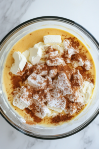 Top-down view of a large glass mixing bowl placed on a white marble countertop, containing cream cheese, butter, powdered sugar, brown sugar, cinnamon, vanilla pudding mix, and lemon juice. A hand mixer is being used to beat the ingredients into a smooth and creamy texture. The pale, fluffy mixture is the rich base for the caramel pecan cheeseball recipe.