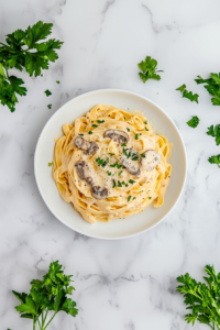 Top-down view of creamy mushroom fettuccine pasta served on a white ceramic plate over a white marble countertop. The pasta is generously coated in a rich cream sauce filled with mushrooms and parmesan, then garnished with fresh chopped parsley. The dish looks elegant, warm, and inviting, perfect for a cozy dinner.