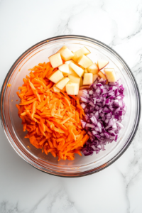 Top-down view showing diced red apple and finely chopped purple onion added to the grated carrots in a clear glass mixing bowl on a white marble countertop. A white cutting board and silver knife rest nearby. These fresh ingredients add a balance of sweetness and sharpness to the vibrant salad base.