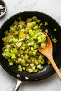 A top-down view of a black skillet placed over a white marble cooktop, filled with melted butter as diced onions and celery cook. The onions are turning translucent, and the celery is softening, releasing a rich aroma. Nearby, small piles of salt, poultry seasoning, and black pepper wait to be added for flavor.
