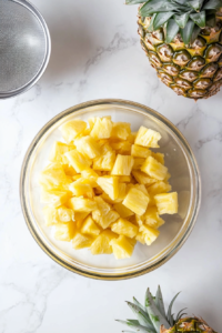 A top-down image of crushed pineapple placed in a clear glass mixing bowl after being drained, with the opened 20-ounce can of pineapple nearby. The setup is on a white marble countertop and signals the first step of the recipe where the canned fruit is prepared for blending with the dry and wet ingredients.