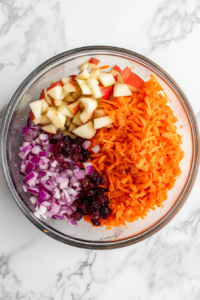 Top-down view of dried cranberries (craisins) being sprinkled into a bowl containing grated carrots, diced apple, and chopped purple onion. The large glass mixing bowl sits on a white marble surface. These dried fruits add a sweet-tart flavor and chewy texture, enhancing the color and taste of the salad.