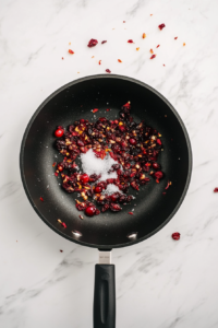 Top-down view of dried cranberries, white sugar, and water being cooked together in a black saucepan on a white marble surface. The cranberries are starting to plump and release their juices. This step allows the berries to soften and develop a sweet base, which will balance the heat from the jalapeños added later.
