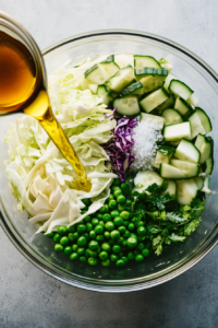 A top-down view of extra virgin olive oil, apple cider vinegar, and white vinegar being drizzled into a large glass bowl of cabbage, cucumbers, green onions, and peas. The white marble surface holds measuring spoons and small bowls with dressings, emphasizing a clean and precise step in flavoring the salad.