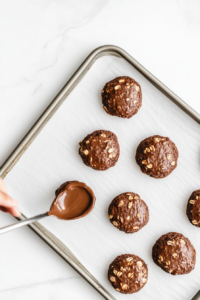 A spoon is used to drop rough scoops of the chocolate oatmeal mixture onto the parchment-lined tray. Each portion is spaced apart for cooling and shaping. The saucepan sits nearby with remaining mixture. This process forms the cookie shapes before they cool and firm up. Everything rests on a white marble surface.