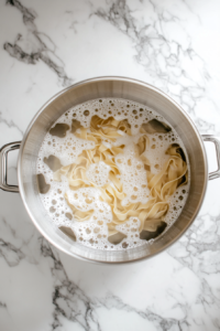Top-down view of a large silver pot filled with boiling salted water, with fettuccine egg pasta cooking inside. The pot is set on a clean white marble countertop, and a wooden spoon rests nearby. The pasta strands are partially submerged and gently swirling in the water. This image captures the beginning step of preparing pumpkin fettuccine, ensuring the pasta is cooked to a perfect al dente texture before being added to the flavorful sauce later on.