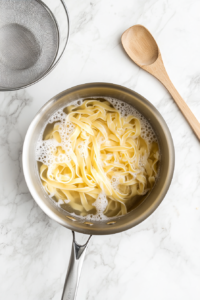 Top-down shot of fettuccine pasta boiling in salted water inside a large stainless steel pot placed on a clean white marble countertop. The pasta is nearly al dente, and the bubbles on the surface show active boiling. This is the first step in preparing the pasta base for the creamy mushroom sauce.